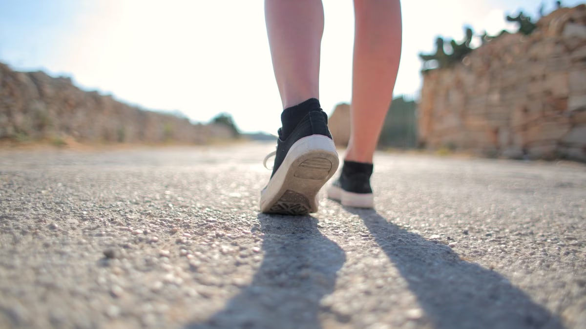 A person walks on a sunny rural road wearing sneakers, casting a long shadow.